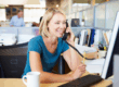 White, blonde woman talks on phone in front of computer monitor in an open office. Background is blurred. A man and other cubicles are seen behind her.