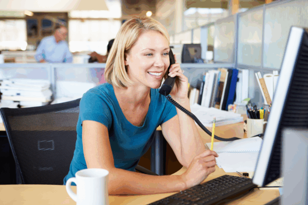 White, blonde woman talks on phone in front of computer monitor in an open office. Background is blurred. A man and other cubicles are seen behind her.