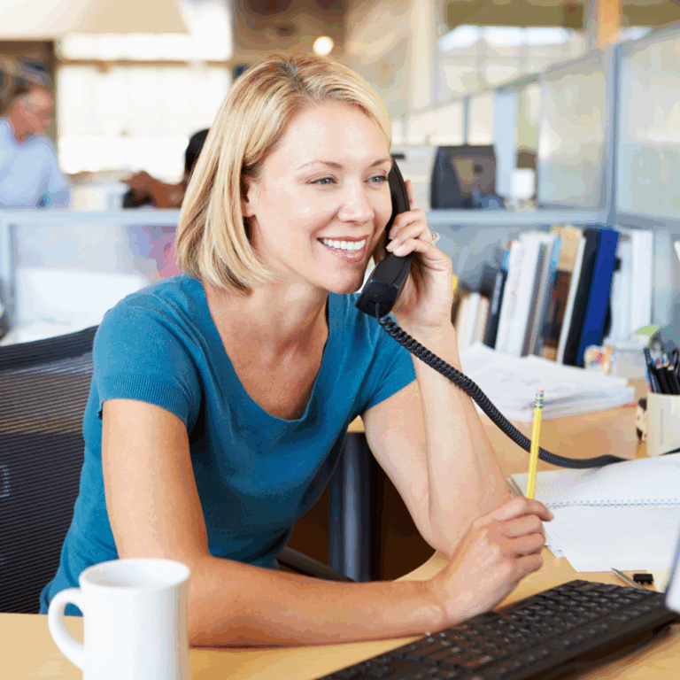 White, blonde woman talks on phone in front of computer monitor in an open office. Background is blurred. A man and other cubicles are seen behind her.