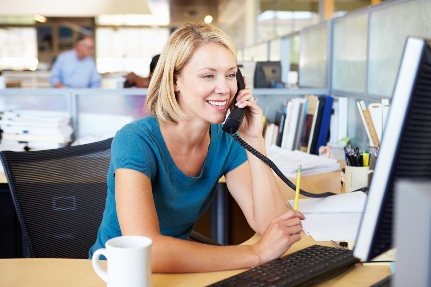 White, blonde woman talks on phone in front of computer monitor in an open office. Background is blurred. A man and other cubicles are seen behind her.