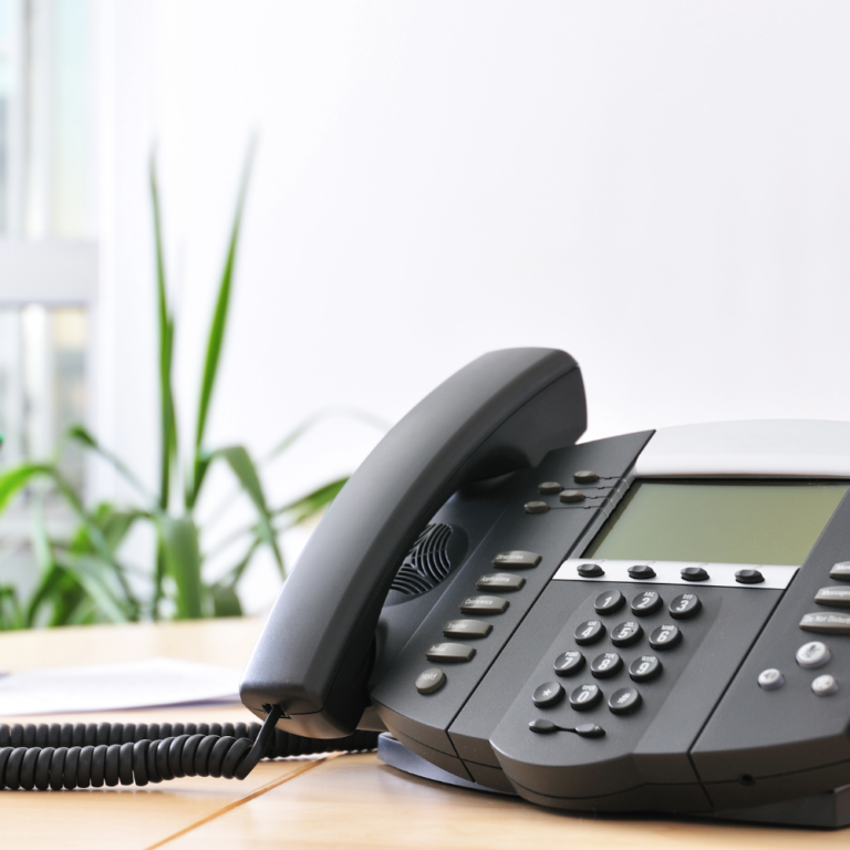 7 Common Mistakes When Choosing a Business Phone System Advanced VoIP business phone system on wooden work desk in office. A cup of markers, a floor plant, and a window are blurred in the background.