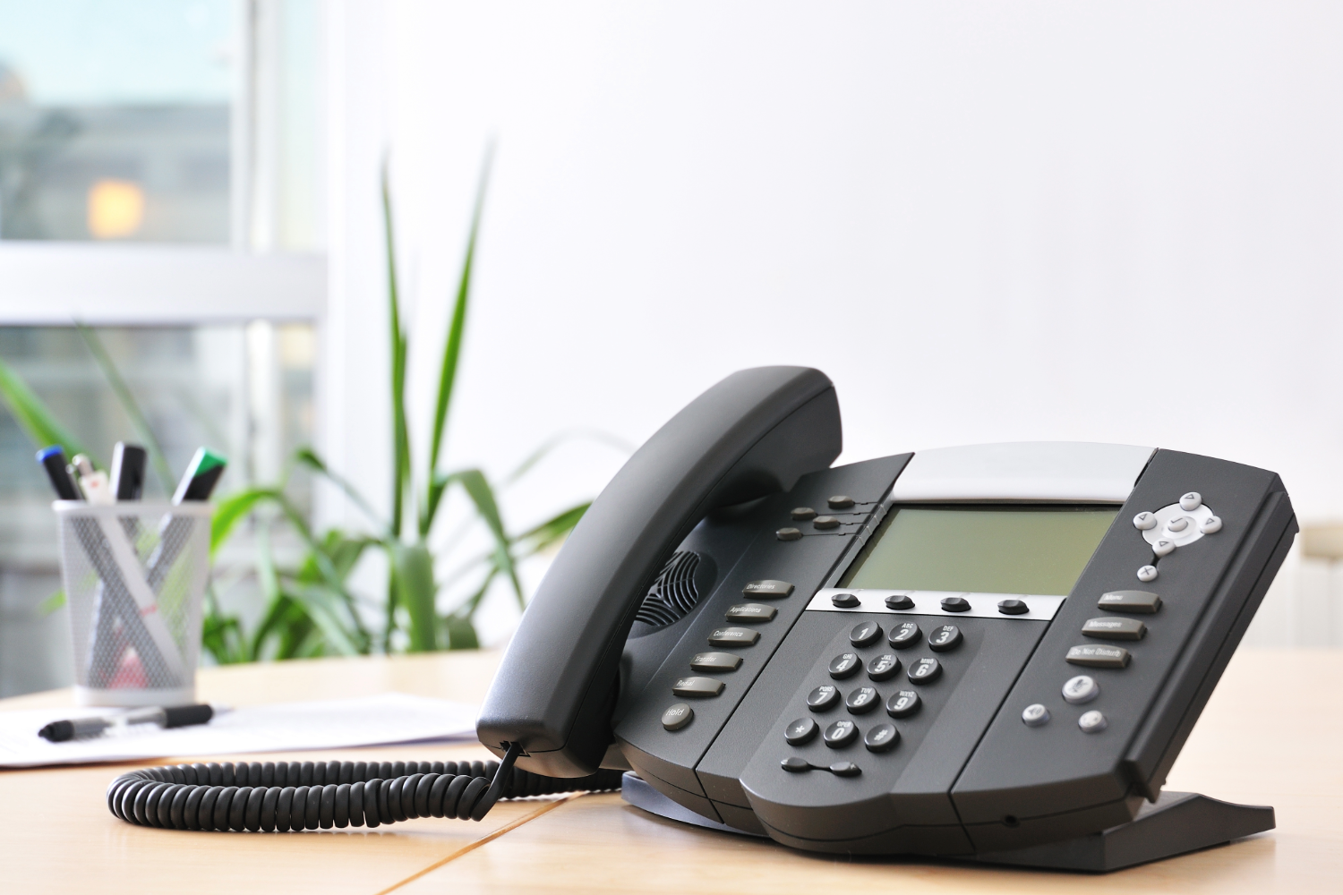 7 Common Mistakes When Choosing a Business Phone System Advanced VoIP business phone system on wooden work desk in office. A cup of markers, a floor plant, and a window are blurred in the background.