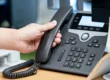 A person's hand lifting the handset off a black office VoIP desk phone